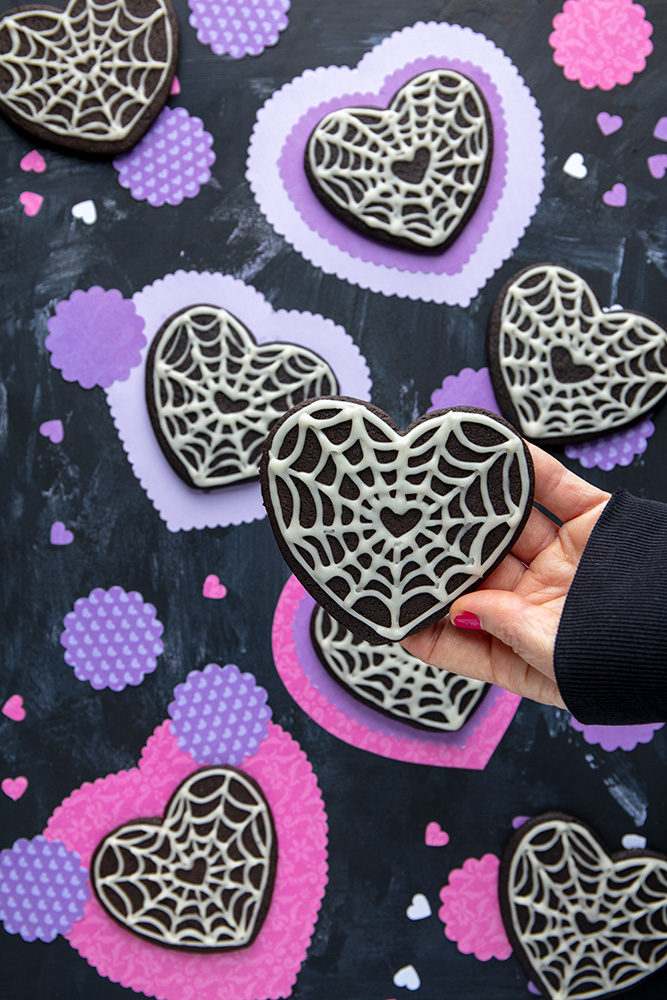 holding a black Halloween heart cookie decorated with spider web icing for goth Valentine’s Day