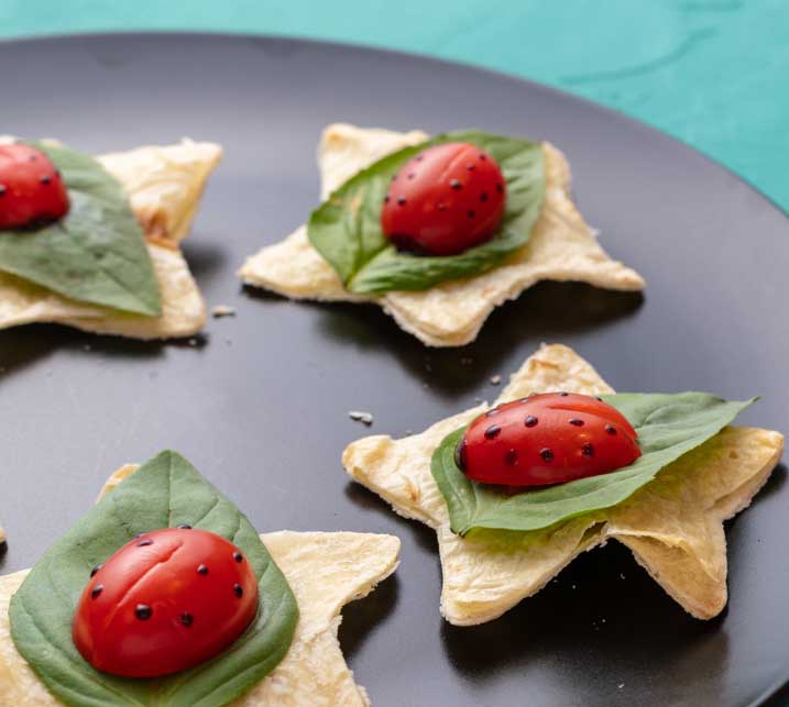 grape tomatoes decorated like ladybugs witting on basil leaves on top of a star-shaped tortilla chip.