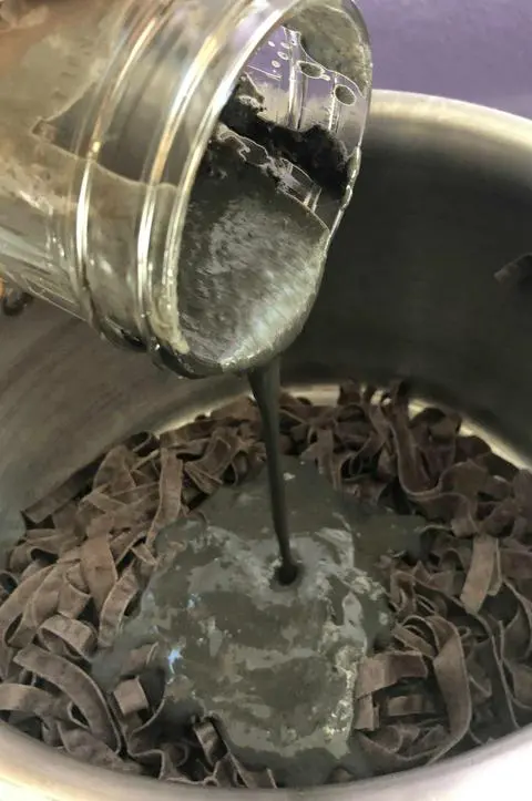 pouring a creamy black sauce over a pot of black wide noodles