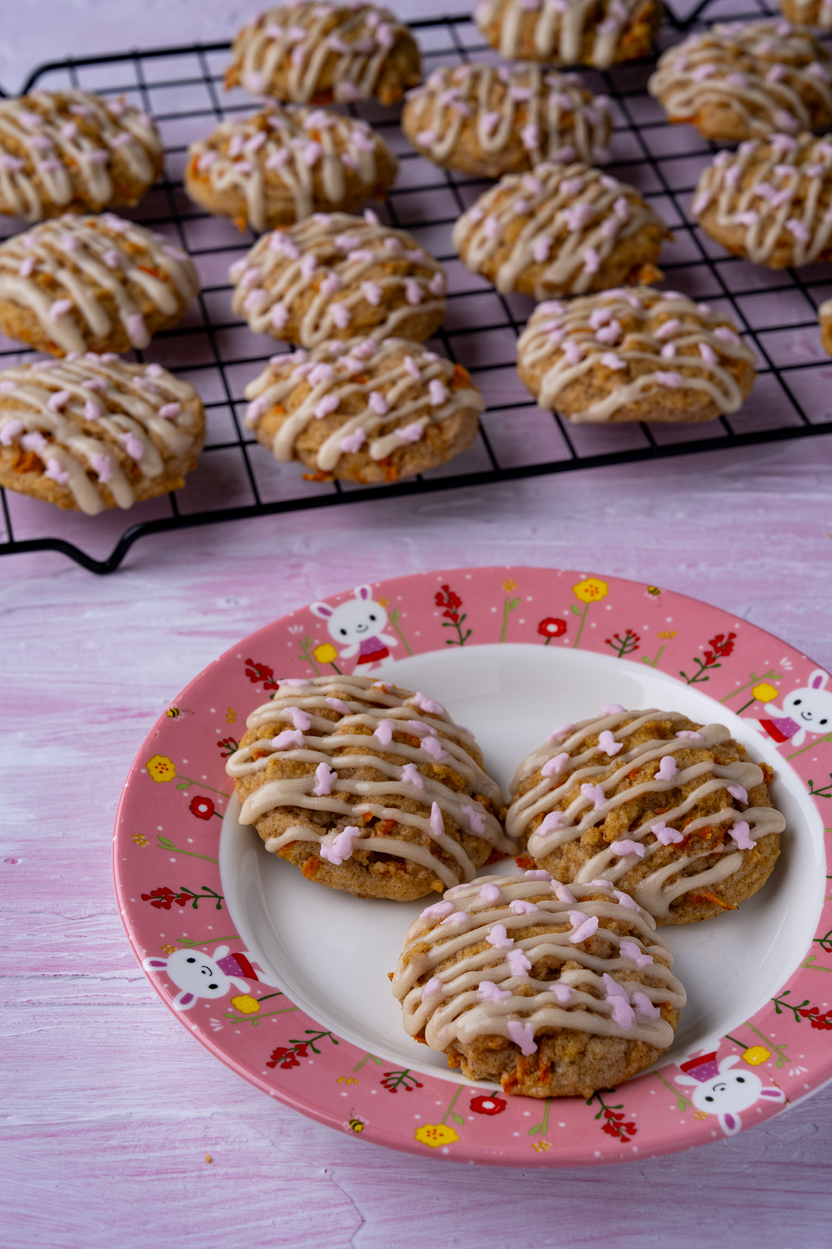 carrot cake cookies with cream cheese icing drizzle and pink bunny sprinkles on a bunny plate with a cooling rack of more cookies behind the plate.