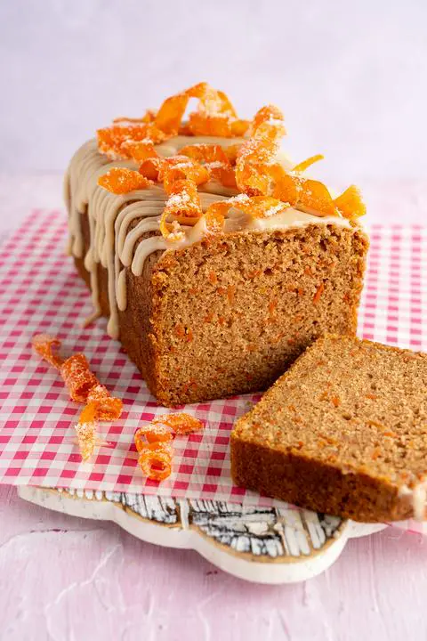 vegan carrot cake loaf with cream cheese icing and candied carrot twists on top and a slice removed.