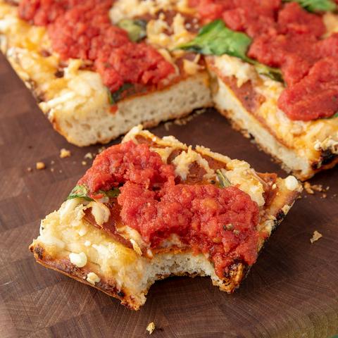 a slice of vegan detroit style pizza with a bite missing and the rest of the pizza behind it