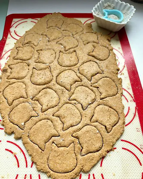 dog treat dough rolled out on a rolling mat with mini cats cut out and a mini cat cookie cutter in a tiny dish of flour.