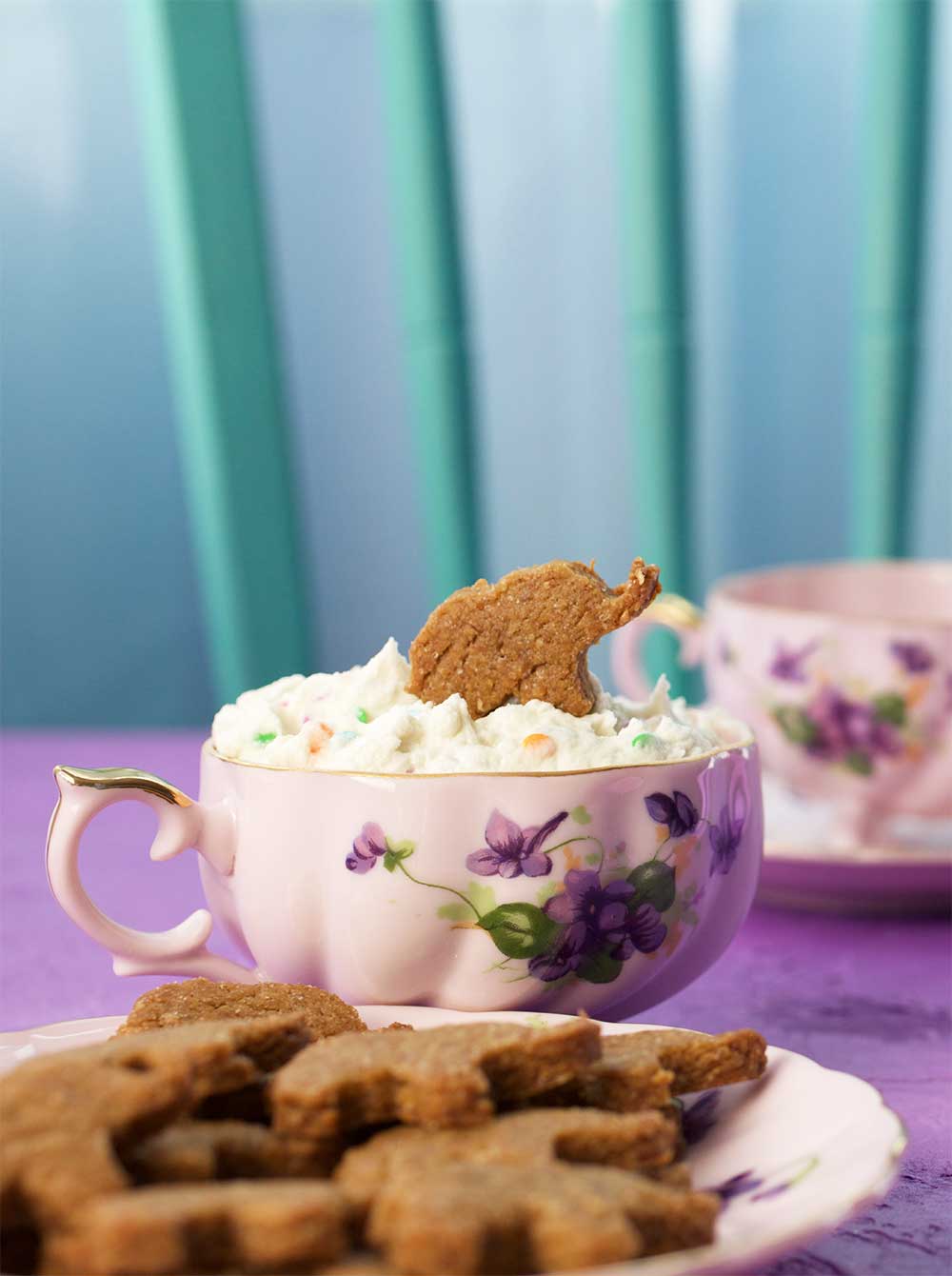 elephant shaped cookie being dipped into a funfetti frosting ina  teacup with a saucer filled with elephant cookies in the foreground.