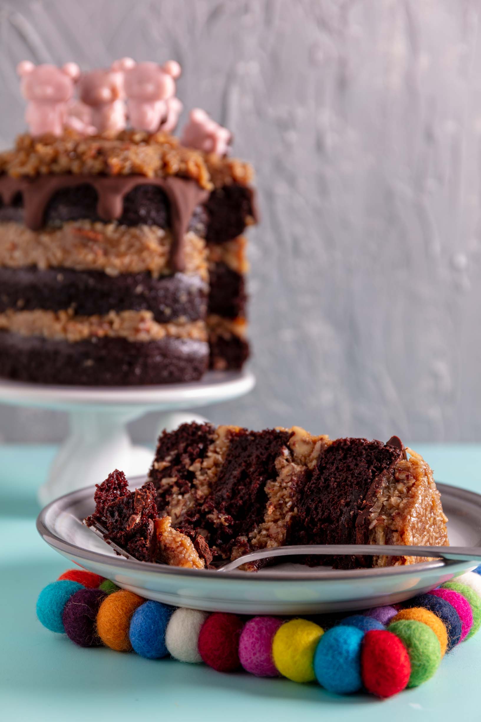 a bite of vegan german chocolate cake on a fork on a plate with a slice of cake with the rest of the cake out of focus in the background