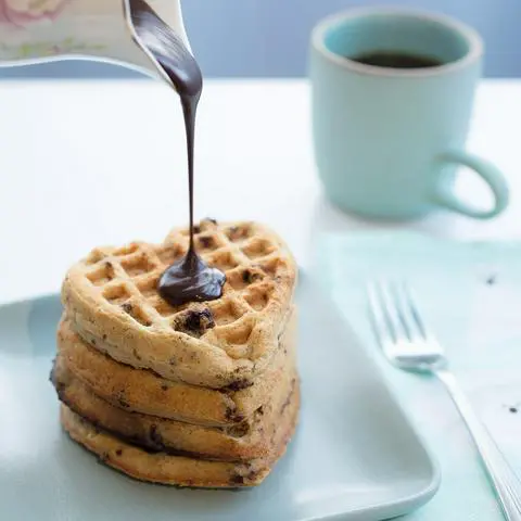 A stack of heart shaped waffles with chocolate syrup being poured on them