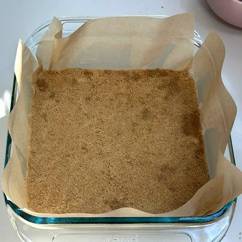 square glass pan lined with parchment paper with a vegan gluten-free packed graham cracker crust at the bottom.