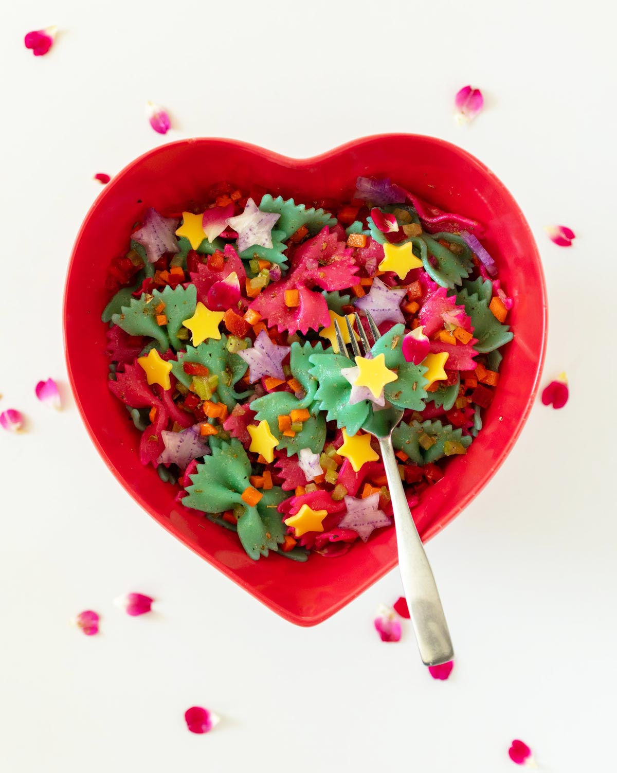 heart shaped bowl filled with multi-colored bowtie shaped pasta and cheese stars and rose petals.