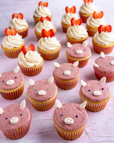 vegan strawberry filled cupcakes decorated in 2 different ways: in the front are pigs and in the back are white frosting with a strawberry heart garnish in the shape of a heart.