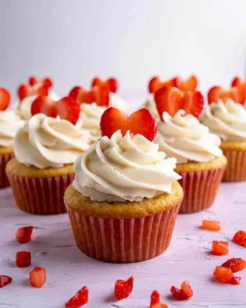 a dozen decorated vegan strawberry cupcakes with strawberry filling scattered around the table.