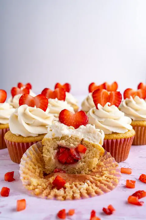 a dozen vegan cupcakes with a bite missing from one showing the fresh strawberry filling inside.