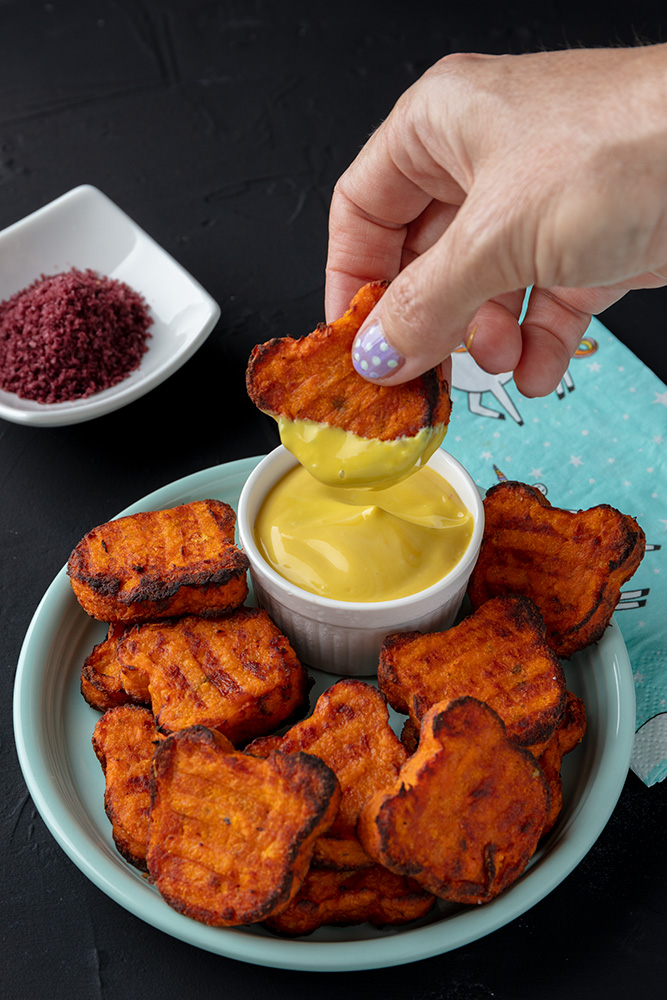 dipping a teddy tot into dip on a plate of bear shaped sweet potato tots, with a dish of purple salt, and a unicorn napkin in the background